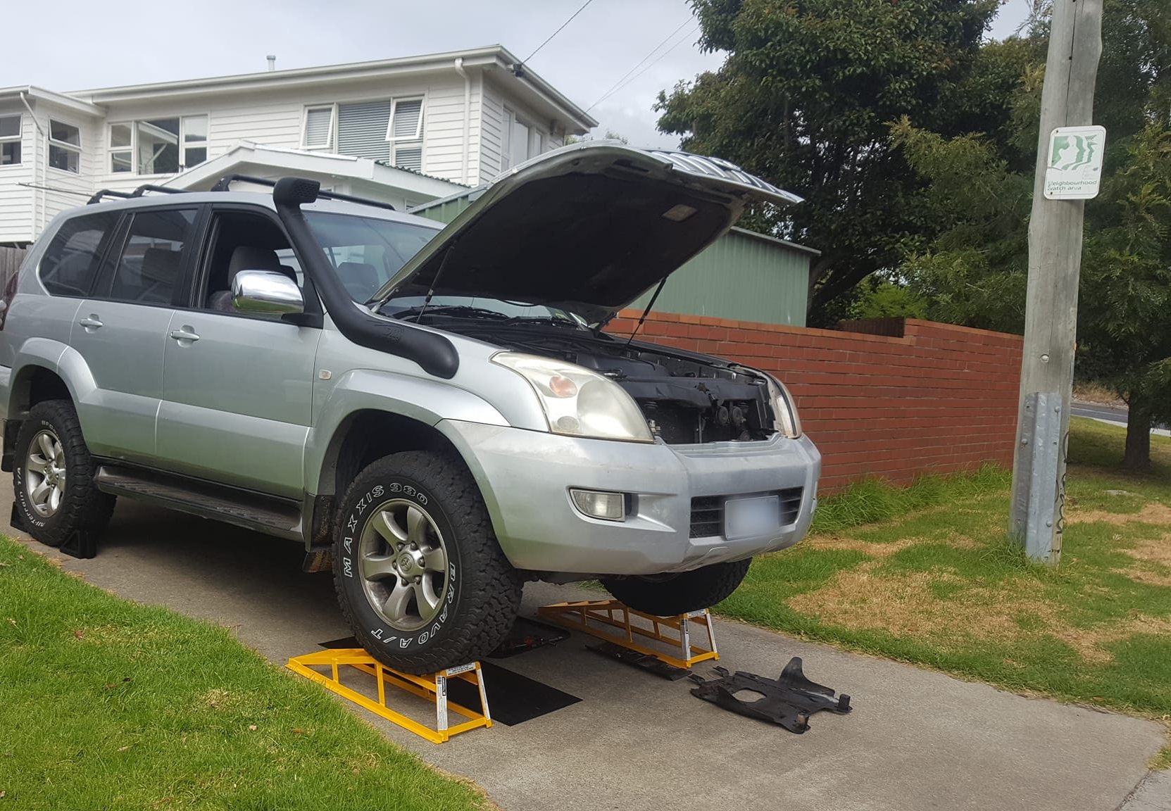 A silver Toyota Prado up on ramps in a suburban driveway with the bonnet open and the bash plate off